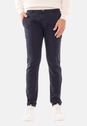 Chino in cotone blu navy con vestibilità slim, caratterizzati da una consistenza liscia, chiusura con bottone, tasche laterali e sneakers bianche per contrasto.
