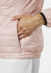 Ljusrosa puffjacka med en ficka med dragkedja på sidan. Den har resår vid fållen och en svart manschettdetalj.