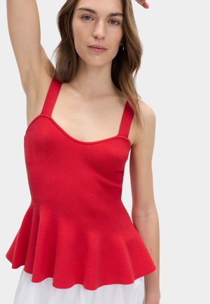 Młoda kobieta w czerwonym bezrękawnym topie peplum z szerokimi ramiączkami, podnosząca jedną rękę, na jednolitym jasnym tle.