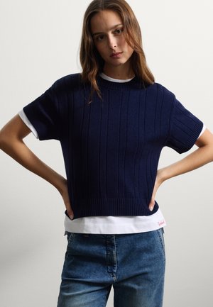 Marineblauwe ribgebreide top met korte mouwen, met een ronde halslijn en een contrasterende witte onderlaag. Gestyled met lichte denim jeans.
