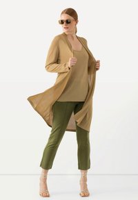 Cardigan long et ouvert en tricot doré, porté sur un débardeur assorti, associé à un pantalon vert olive et des sandales métalliques à lanières.