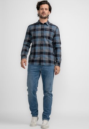 Blauwe en grijze geruite flanellen shirt met lange mouwen, knoopsluiting aan de voorkant en twee borstzakken, gecombineerd met lichtblauwe spijkerbroek en witte sneakers.