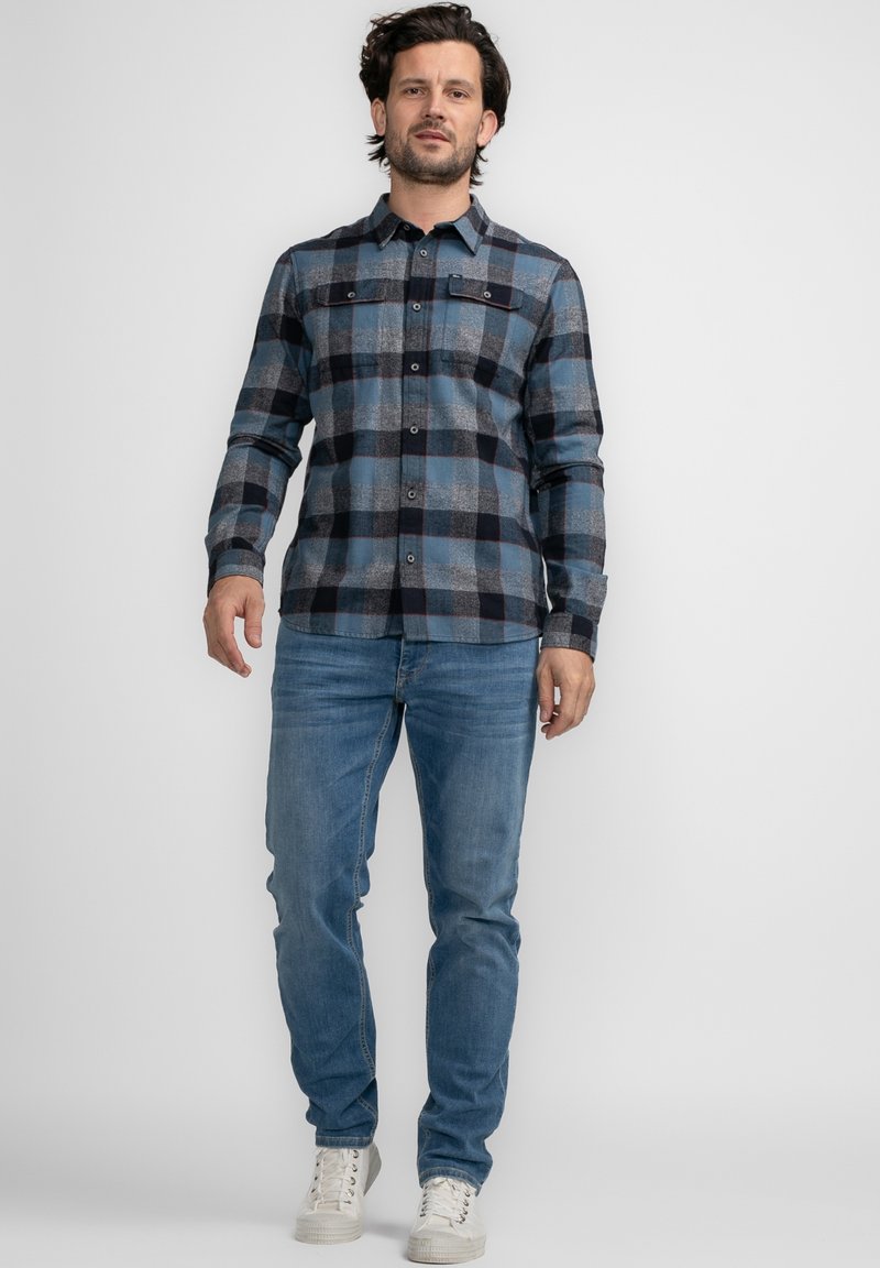 Camisa de franela a cuadros azul y gris con mangas largas, botonadura frontal y dos bolsillos en el pecho, combinada con jeans azul claro y zapatillas blancas.