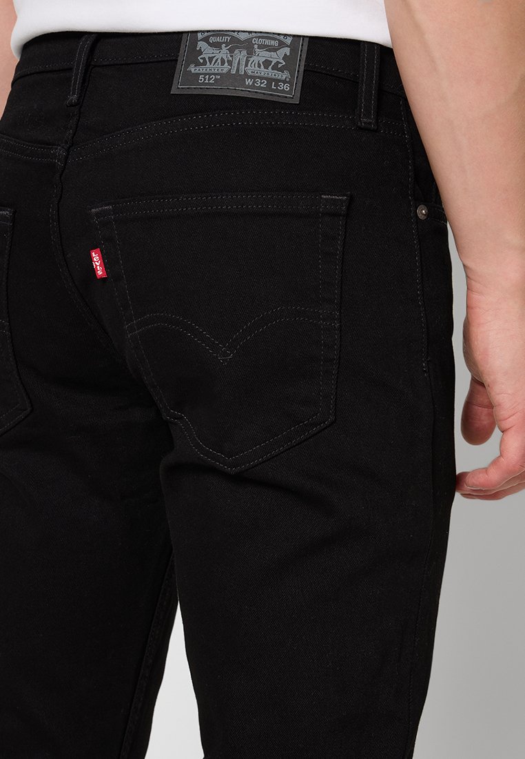 Black denim jeans with rear pockets, contrasting stitching, and a black leather patch featuring brand logo and details on the waistband.
