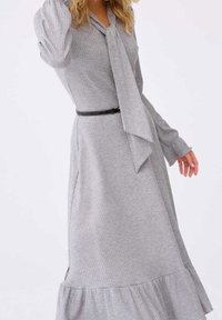 Femme portant une robe grise côtelée à manches longues avec une ceinture noire et un détail d'attache de cou assorti.