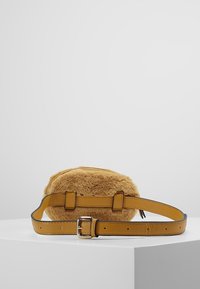 Kulatá taška na opasek z umělé kožušiny se zeleným páskem a stříbrnou sponou položená na bílé ploše.