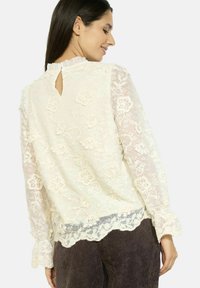 Blusa bianca in pizzo con maniche lunghe trasparenti, scollatura arricciata e apertura a chiave sul retro. Motivi floreali decorativi e orlo a zig-zag.