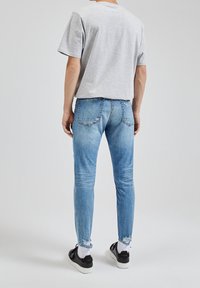 Grå kortärmad skjorta, ljusblå slitna skinny jeans, svarta skor och vita strumpor, sedd bakifrån mot en enkel bakgrund.