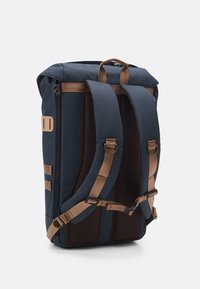 Sac à dos bleu marine avec bretelles marron ajustables et rembourrées, et détails en mesh. Dispose d'une poignée supérieure et d'une fermeture éclair latérale. Tissu léger.