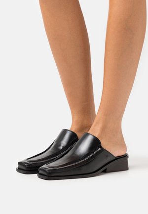 Mules de piel negra sin cordones con punta afilada, detalle de costura blanca y un pequeño tacón cuadrado. Textura suave y diseño minimalista.