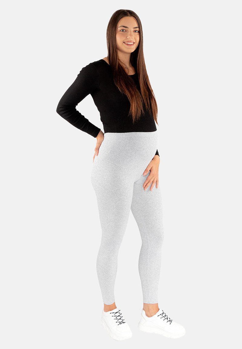 Szare legginsy wykonane z elastycznego materiału, połączone z czarną bluzką z długim rękawem i białymi sneakersami. Modelka stoi z jedną ręką na biodrze.