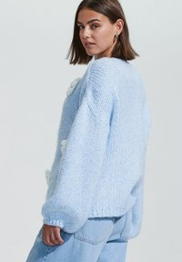 Une femme aux cheveux bruns mi-longs pose de profil, portant un pull en tricot bleu clair orné de petites fleurs blanches.