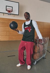 Fekete Chicago Bulls ujjatlan felső fehér hosszú ujjú póló felett, piros bő nadrág, fehér sportcipő, fekete kosárlabdával a kézben és egy drótkosár kosárlabdákkal.