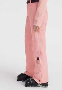 Pink snow pants with a relaxed fit, featuring a textured fabric, side pocket, and black accents. Worn with black ski boots.