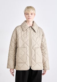 Καμηλό καπιτονέ μπουφάν με φαρδύ γιακά, μπροστινό φερμουάρ και δύο μεγάλες μπροστινές τσέπες. Το διαμαντέ σχέδιο στο ύφασμα προσθέτει υφή.