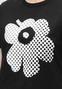 Μαύρο T-shirt με μεγάλο λευκό σχέδιο λουλουδιού από μαύρες πουά κουκκίδες διαφόρων μεγεθών που καλύπτουν τα πέταλα και το κέντρο.