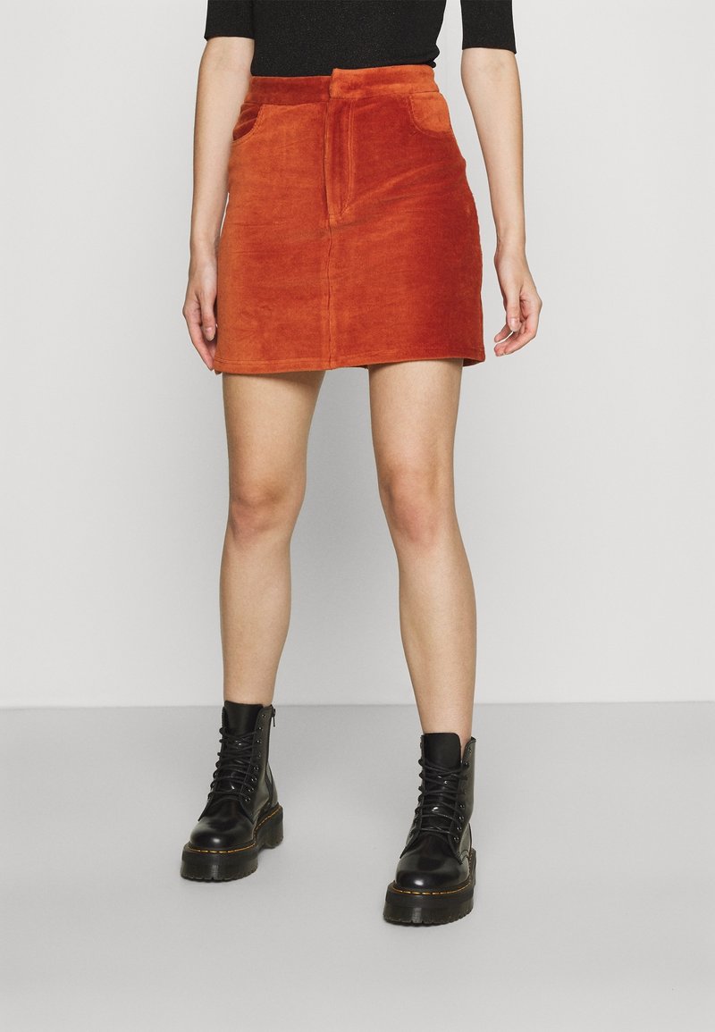 Mini-jupe en velours côtelé couleur rouille avec poches avant et fermeture éclair, associée à des bottes de combat noires à lacets. Texture lisse.