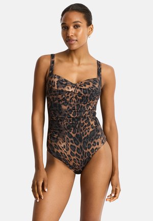 Mujer con un bañador entero de estampado de leopardo marrón y negro con tirantes anchos, de pie contra un fondo liso y claro.