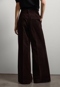 Bruine corduroy wijdere broeken met achterzakken, hoge taille en verticale strepen, weergegeven van achteren tegen een neutrale achtergrond.