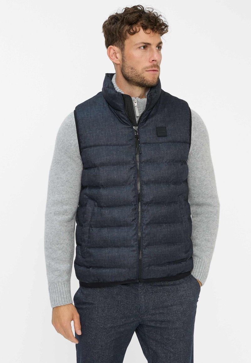 Dicker Weste aus dunkelblauem Stoff mit hohem Kragen und Reißverschluss vorne. Getragen über einem hellgrauen Pullover, mit einem Logo-Patch auf der Brust.