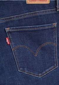 Jeans de mezclilla azul oscuro con un bolsillo trasero que presenta costuras amarillas en contraste, un diseño curvado y una etiqueta roja de Levi's cerca del borde del bolsillo.