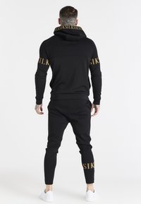 Conjunto de sudadera con capucha negra y pantalones jogger, con acentos de logo dorados, diseño ajustado y una tela texturizada, ideal para el uso casual.