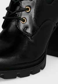 Gros plan d'une chaussure noire en cuir brillant à lacets avec œillets dorés et semelle épaisse et robuste.