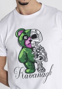 Bijela majica s grafičkim dizajnom podijeljenog medvjeda – jedan dio je zeleni pliš, a drugi dio je robotski kostur. Ispod je ime "Kavanagh."