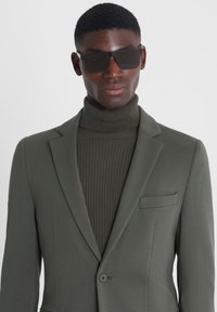 Chaqueta de traje verde oliva con solapas estructuradas y un bolsillo en el pecho, combinada con un jersey de cuello alto de canalé oscuro y gafas de sol rectangulares negras.
