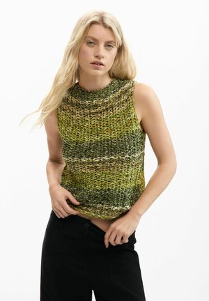 Donna con lunghi capelli biondi che indossa un top verde e marrone senza maniche in maglia e pantaloni neri su uno sfondo semplice.