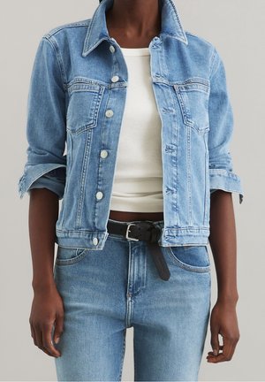 Osoba nosząca jasnoniebieską dżinsową kurtkę z podwiniętymi rękawami, biały krótki top, jasnoniebieskie dżinsy oraz czarny pasek z srebrną klamrą.