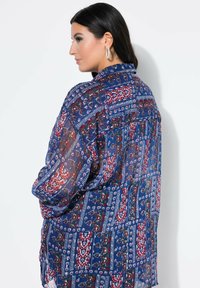 Una mujer con cabello oscuro lleva grandes aretes de aro y una camisa azul transparente con patrón, de perfil contra un fondo blanco.