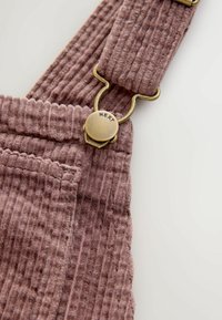 Lyserøde corduroy overalls med teksturerede lodrette ribber, en messingklip med mærket "NEXT" og en lomme detalje på stropen.