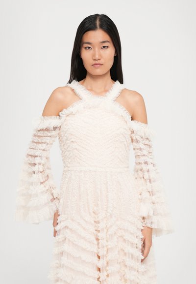 Vrouw met steil zwart haar, gekleed in een witte off-shoulder jurk met doorschijnende rimpelige lange mouwen tegen een effen achtergrond.