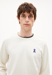Jonge man met kort bruin haar, gekleed in een witte blouse met lange mouwen en een klein donkergpaars symbool op de borst, staande tegen een effen achtergrond.