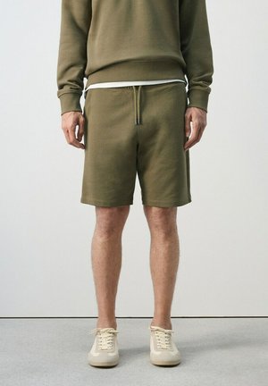 Hombre con sudadera verde oliva y pantalones cortos a juego con zapatillas beige, de pie contra un fondo claro liso.