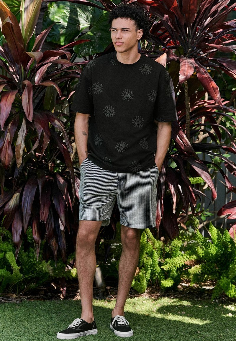 Černé tričko s krátkým rukávem a vzorem slunečního paprsku, doplněné o šedobílé pruhované šortky. Černé tenisky dotvářejí outfit.