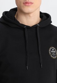 Felpa nera con cappuccio e cordoncino, caratterizzata da una patch circolare del logo Vans sul petto. Il tessuto appare morbido con una texture liscia.