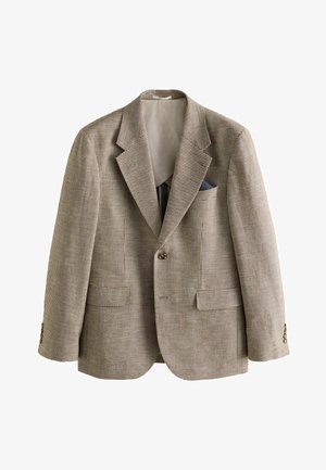 Mann trägt einen braun-schwarz karierten Anzug mit olivgrünem Hemd, steht vor einer schlichten, hellen Wand.