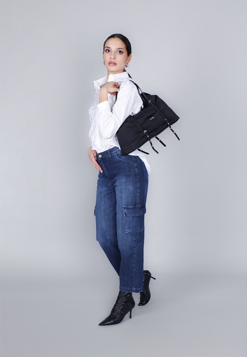 Zwarte stoffen handtas met meerdere ritsen en banden, gecombineerd met een witte blouse met kraag, blauwe cargobroek en spitse zwarte hakken.