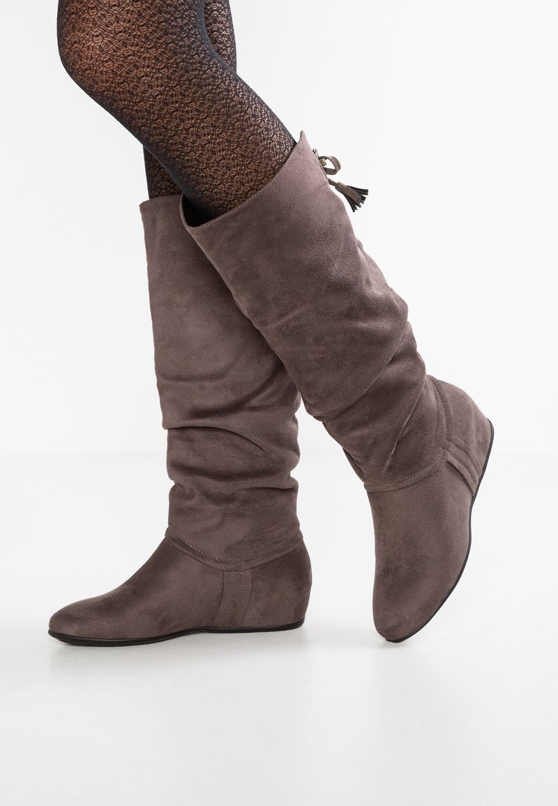 Bottes mi-mollet en daim marron clair portées avec des collants noirs à motifs sur fond blanc.