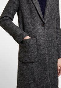 Cappotto in misto lana di colore grigio scuro, con rever a punta, chiusura a un solo bottone e ampie tasche frontali. Finitura testurizzata, vestibilità su misura.