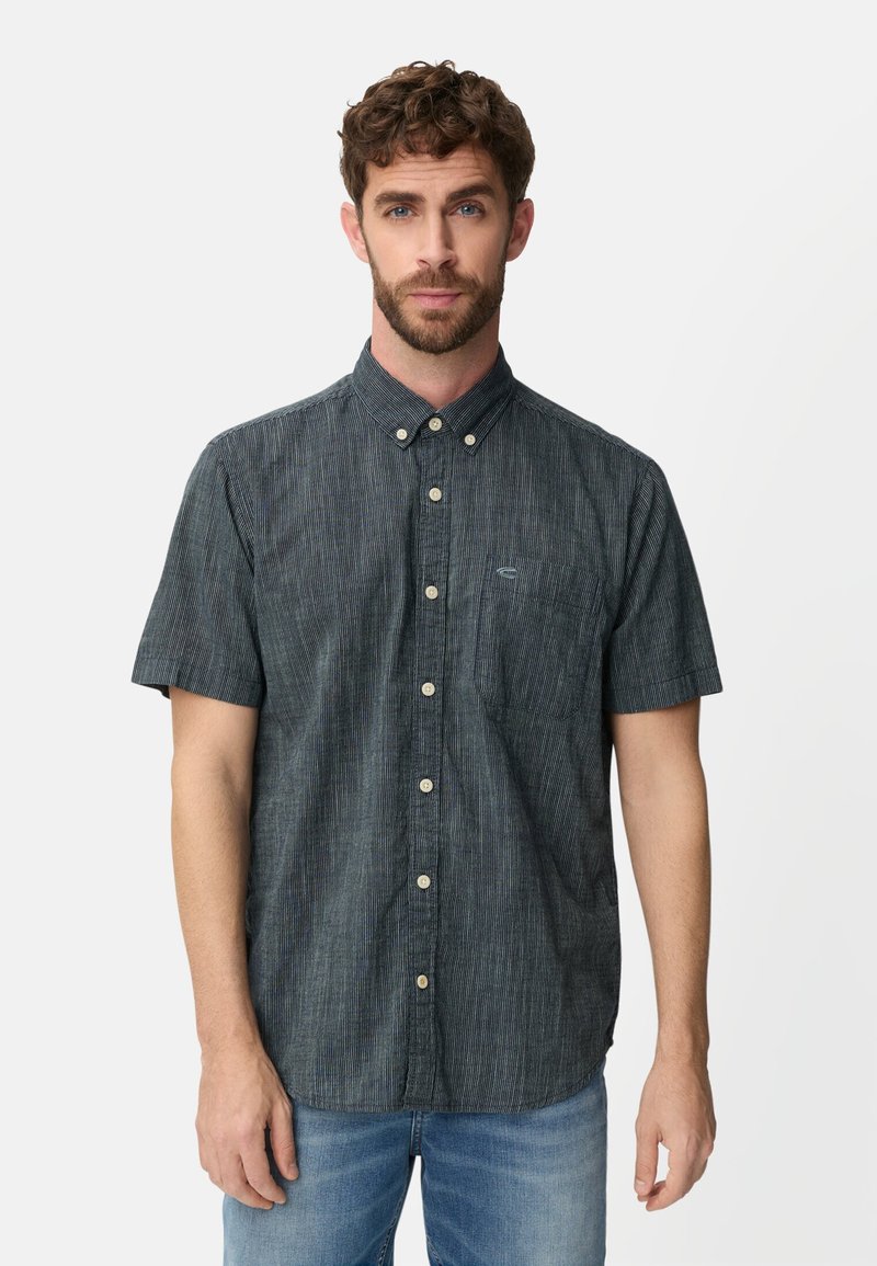 Man met krullend haar en baard, gekleed in een donkergrijs overhemd met korte mouwen en lichtblauwe spijkerbroek, kijkt naar voren tegen een witte achtergrond.