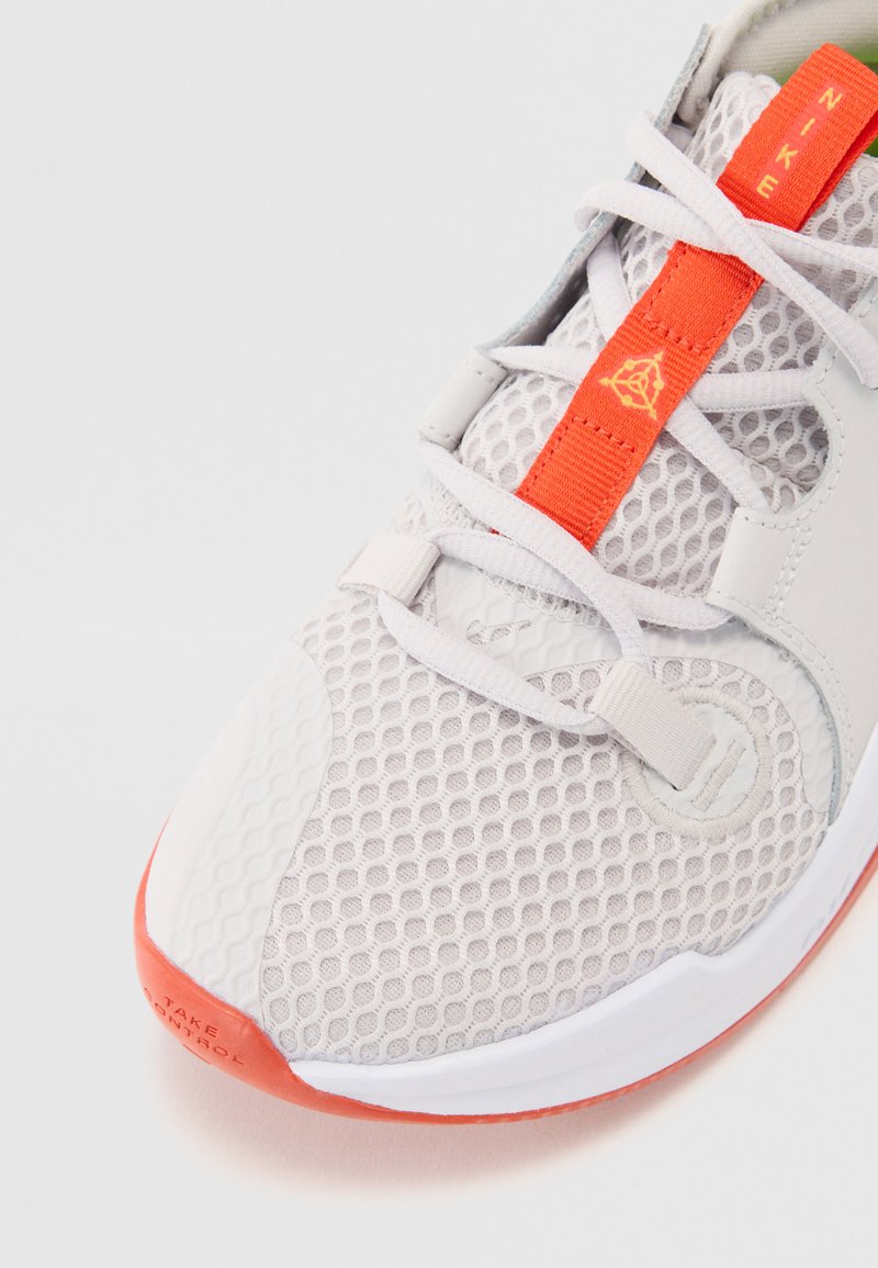 Zapatilla deportiva gris claro con parte superior de malla, acentos naranjas y suela de goma texturizada. Cuenta con cordones ajustables y detalles del logo.