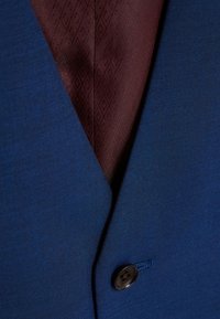 Veste de costume bleue en tissu texturé, dotée d'une doublure bordeaux foncé avec un motif en diamants et d'une fermeture à un seul bouton.