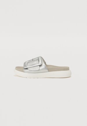 Sølv slip-on sandal med stor spænde rem og hvid polstret sål på en ensartet lys baggrund.