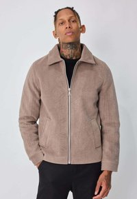 Veste taupe zippée en tissu doux et texturé. Caractérisée par un col pointu et des poches latérales. Portée par-dessus un haut noir, design ajusté.