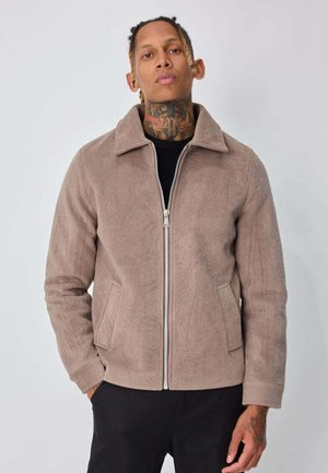 Veste taupe zippée en tissu doux et texturé. Caractérisée par un col pointu et des poches latérales. Portée par-dessus un haut noir, design ajusté.