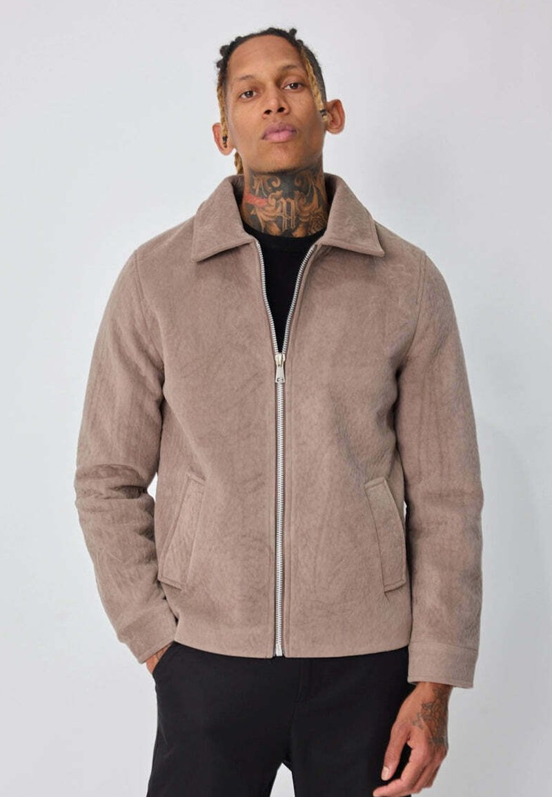 Veste taupe zippée en tissu doux et texturé. Caractérisée par un col pointu et des poches latérales. Portée par-dessus un haut noir, design ajusté.