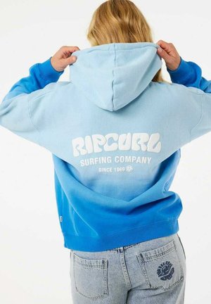 Osoba w niebieskiej gradientowej bluzie Ripcurl z kapturem i jasnoniebieskich dżinsach, trzyma kaptur, plecami do widza, pokazując logo i tekst na bluzie.